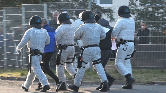 Polizisten in weißer Schutzkleidung mit schwarzen Helmen und schwarzen Stiefeln führen zwei Männer ab